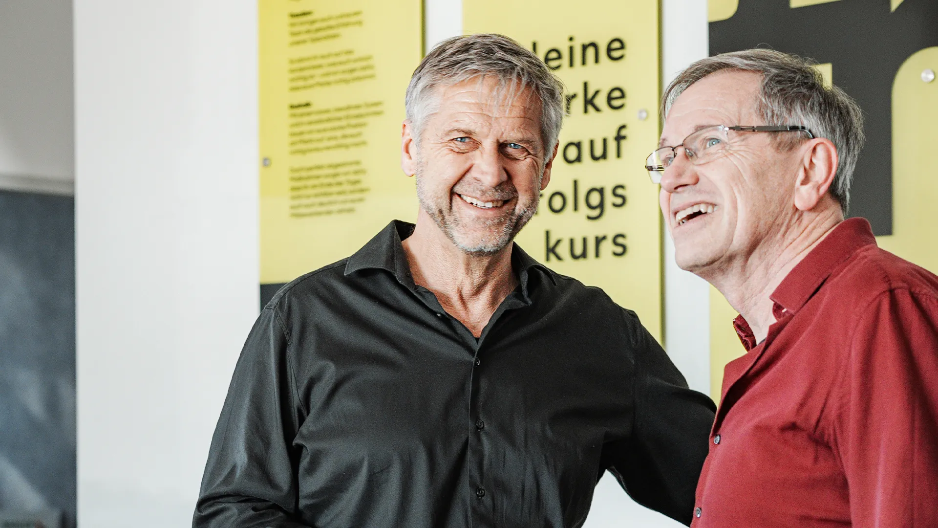 Zwei Personen lachen gemeinsam in einem Gespräch, im Hintergrund eine Wand mit Text