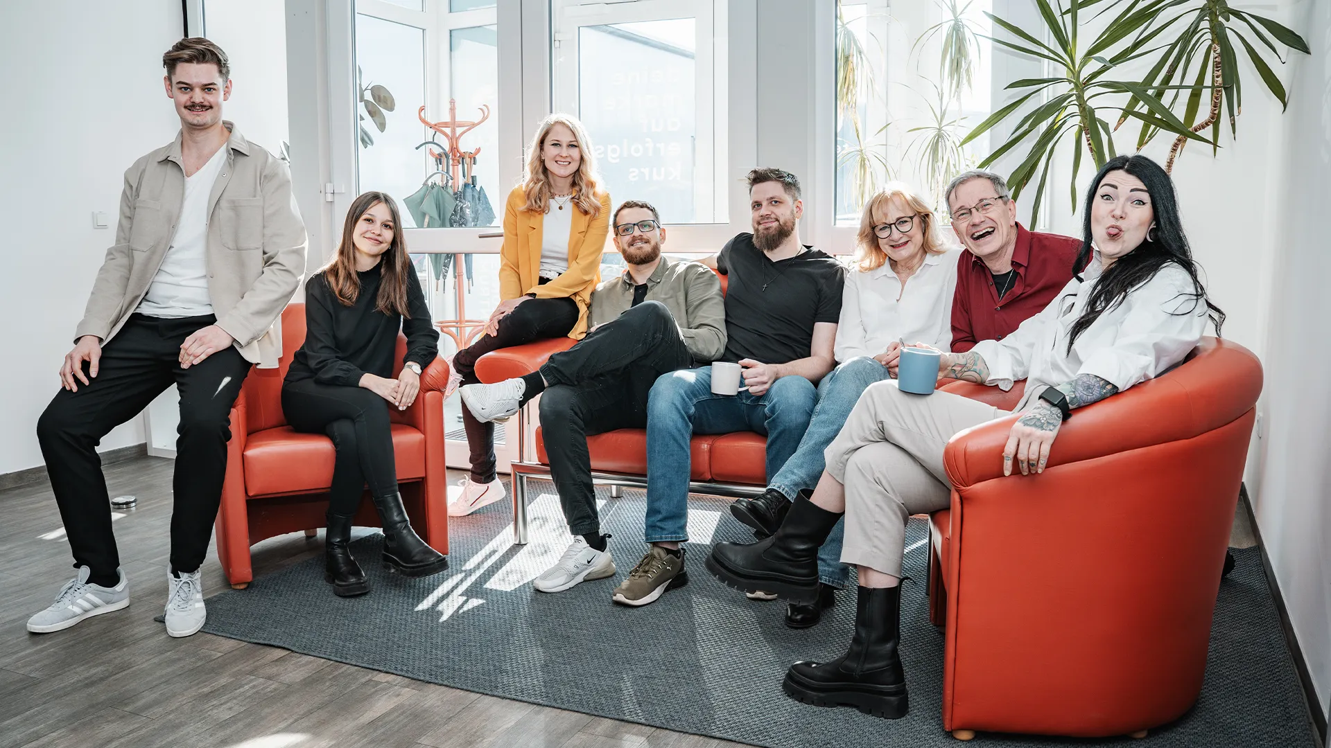 Alle neun Team Marke Mitglieder sitzen auf einer roten Couch und lachen in de Kamera